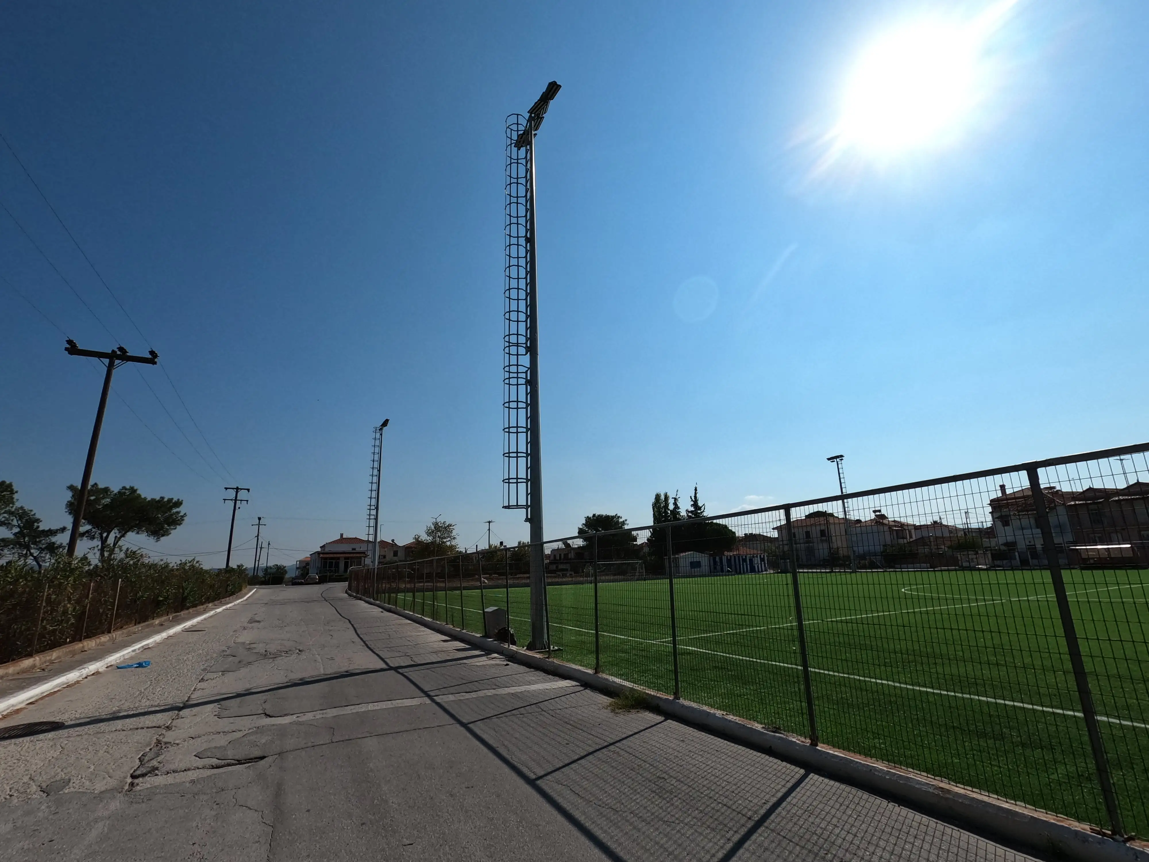 Φάινεται λεπτομέρεια ο φράχτης του γηπέδου και η σκάλα από τον ένα ιστό φωτισμού. Παράλληλα με τον φράχτη φαίνεται και ο δρόμος.